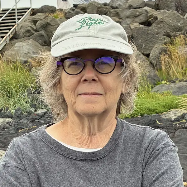 Janie Braverman in gray shirt and baseball cap, standing in front of a rock-filled bank.